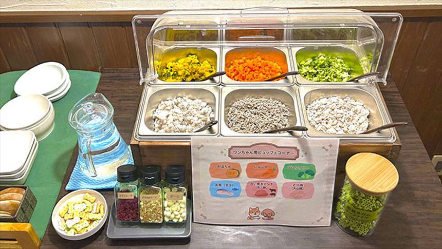鳥取県・大山の犬と泊まれる宿「ホテル旬香大山リゾート」|「ワンちゃん用ビュッフェコーナー」と書かれた木製の台。手前にビュッフェメニュー(かぼちゃ、にんじん、ブロッコリー、お魚、合挽きとシカ、とり肉)をイラストで紹介するPOPがあり、その奥にガラスケースに入ったビュッフェ料理が並んでいる。