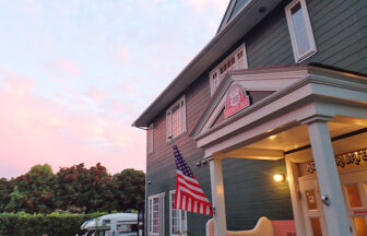 夕焼けに染まる空の下、アメリカ国旗が揺れる洋風建築の宿泊施設の外観。緑の外壁と白い柱が特徴的で、リゾート感のある雰囲気を演出している｜犬と泊まれる静岡県伊豆高原の宿｜古き良きアメリカに憧れてDIYで仕上げた、愛犬と泊まれる全5室のペンション｜ころぶきベース