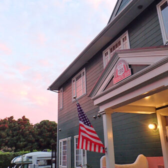夕焼けに染まる空の下、アメリカ国旗が揺れる洋風建築の宿泊施設の外観。緑の外壁と白い柱が特徴的で、リゾート感のある雰囲気を演出している｜犬と泊まれる静岡県伊豆高原の宿｜古き良きアメリカに憧れてDIYで仕上げた、愛犬と泊まれる全5室のペンション｜ころぶきベース