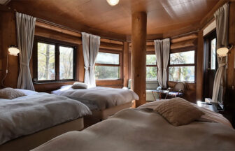 長野県・白馬の犬と泊まれる宿「ログホテル　三―ティア」｜円形の壁面に沿って配置された3台のシングルベッドがある宿泊施設のログルーム。壁と天井はすべて木製で、中央に象徴的な太い木の柱が立っている。各窓には白いカーテンがかかり、枕元にはアンティーク調のランプが設置されている。奥には小さなテーブルと椅子があり、全体的にブラウンとグレーを基調とした落ち着いた色調