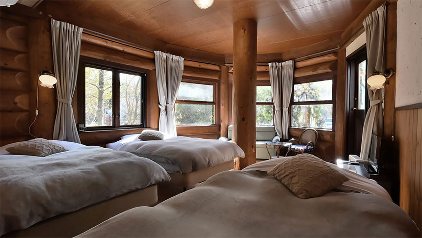 長野県・白馬の犬と泊まれる宿「ログホテル　三―ティア」｜円形の壁面に沿って配置された3台のシングルベッドがある宿泊施設のログルーム。壁と天井はすべて木製で、中央に象徴的な太い木の柱が立っている。各窓には白いカーテンがかかり、枕元にはアンティーク調のランプが設置されている。奥には小さなテーブルと椅子があり、全体的にブラウンとグレーを基調とした落ち着いた色調