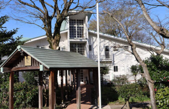 鳥取県・大山の犬と泊まれる宿「ホテル旬香大山リゾート」｜青空の下に佇む、白い外壁と大きな窓が特徴的な3階建ての建物（ホテル・ペンション）。手前には木造の屋根付きエントランスアプローチがあり、周囲を大きな木々や植栽が囲んでいる