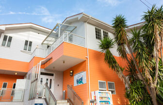 神奈川県の犬と泊まれる宿「CARO FORESTA 城ヶ崎海岸 BASE」｜青空の下に映える、鮮やかなオレンジ色と白の外壁が特徴的な「CARO FORESTA 城ヶ崎海岸 BASE」の外観｜南国風の植物が植えられ、明るく開放的なエントランス。