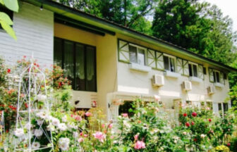 ペンションブルーポピー｜満開のイングリッシュガーデンに囲まれた、2階建ての白いペンションの外観。ピンクや白のバラが咲き誇り、奥には緑豊かな森が見える。