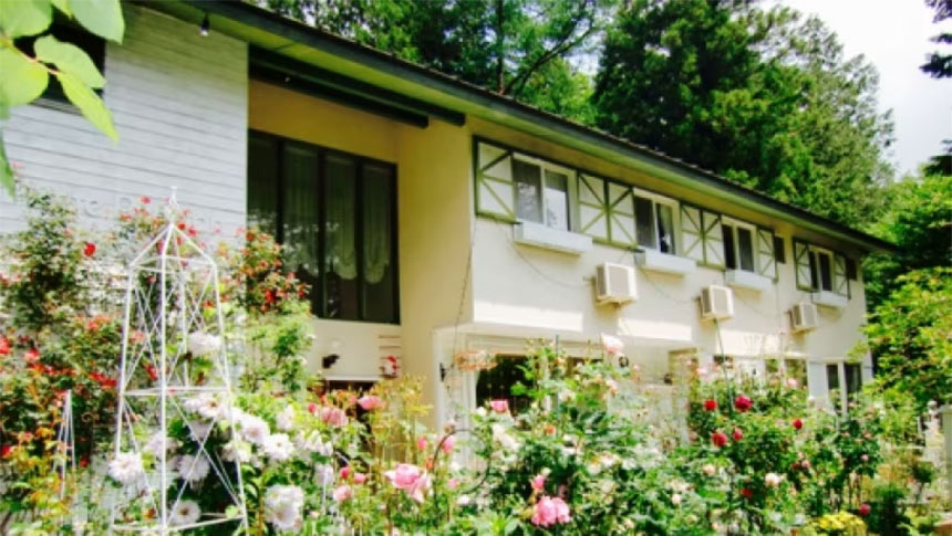 ペンションブルーポピー｜満開のイングリッシュガーデンに囲まれた、2階建ての白いペンションの外観。ピンクや白のバラが咲き誇り、奥には緑豊かな森が見える。