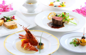 軽井沢ホテルそよかぜ｜白いテーブルに並べられた、ロブスターのソテー、牛フィレ肉のステーキ、フォアグラのパテ、そしてデザートが盛られたフランス料理のフルコース。両脇には鮮やかなピンク色の洋ランの花が添えられている