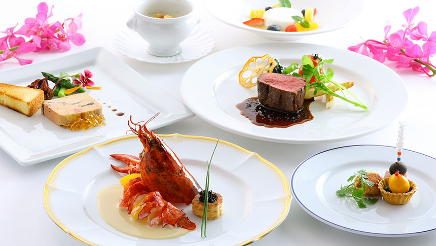 軽井沢ホテルそよかぜ｜白いテーブルに並べられた、ロブスターのソテー、牛フィレ肉のステーキ、フォアグラのパテ、そしてデザートが盛られたフランス料理のフルコース。両脇には鮮やかなピンク色の洋ランの花が添えられている
