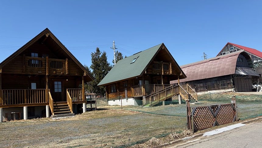 フレンドハウスひるがの｜澄み切った青空が広がる日、木の温かみが感じられる2棟のコテージが並んでいる。手前には、手入れされた砂利道と木のラティスフェンスがある