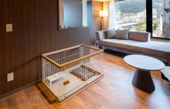 熱海温泉　渚館｜モダンな木目調の壁とフローリングの客室。窓際にはゆったりとしたグレーのソファがあり、手前にはワンちゃん用のケージが設置されている。ケージの中にはトイレシート用のトレーと食事用のスタンドが用意されている。