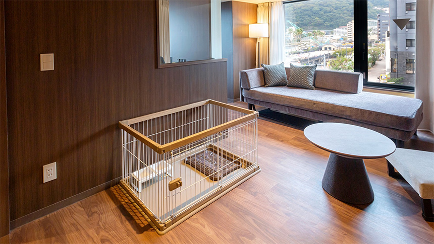 熱海温泉　渚館｜モダンな木目調の壁とフローリングの客室。窓際にはゆったりとしたグレーのソファがあり、手前にはワンちゃん用のケージが設置されている。ケージの中にはトイレシート用のトレーと食事用のスタンドが用意されている。
