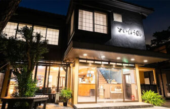 夜空に映える、和風旅館「すいほう園」の温かな光。黒を基調としたシックな外観と、障子窓が落ち着いた雰囲気を醸し出す。玄関先には、宿泊客を迎え入れるように光が灯っている様子