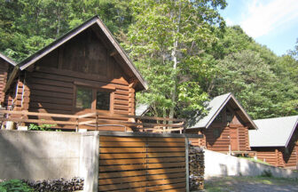 ログコテージさくら｜澄み切った青空の下、鮮やかな緑の木々に囲まれたログハウス。手前には、手入れされた砂利道と薪棚がある