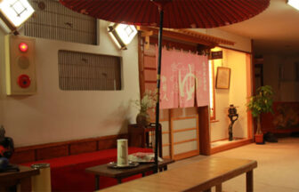 山形県の癒しの源泉かけ流しの宿　温海荘｜和風の情緒あふれる温泉宿のロビー。大きな赤い野点傘が広がり、奥にはピンク色の暖簾がかかった『温泉』の入り口が見える。手前には、赤いフェルトのベンチや湯茶サービスのセットが置かれている