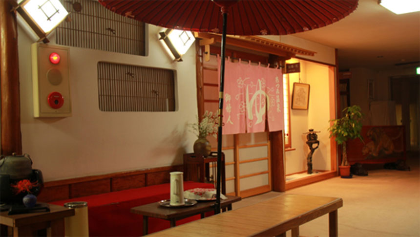 山形県の癒しの源泉かけ流しの宿　温海荘｜和風の情緒あふれる温泉宿のロビー。大きな赤い野点傘が広がり、奥にはピンク色の暖簾がかかった『温泉』の入り口が見える。手前には、赤いフェルトのベンチや湯茶サービスのセットが置かれている