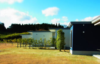 大分県のごーげんばら久住｜緑豊かな山と広々とした芝生を背景に建つ、モダンな青と白の外壁の平屋建て別荘。手前には若い植栽が並んでいる。
