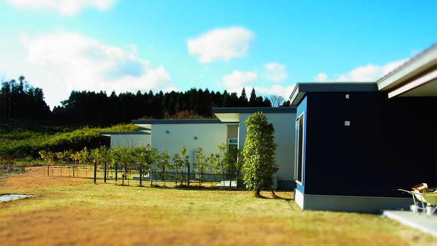 大分県のごーげんばら久住｜緑豊かな山と広々とした芝生を背景に建つ、モダンな青と白の外壁の平屋建て別荘。手前には若い植栽が並んでいる。