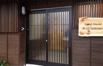 和モダンな趣の「Guest House 楽旅 NOMACHI」の玄関。濃い木目の外壁に、黒い格子戸が映える。右側には施設名が刻まれた木製の看板が掛けられている。