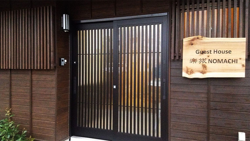 和モダンな趣の「Guest House 楽旅 NOMACHI」の玄関。濃い木目の外壁に、黒い格子戸が映える。右側には施設名が刻まれた木製の看板が掛けられている。