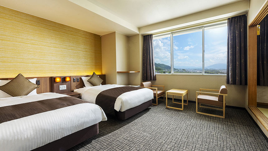 明るい陽光が差し込むツインタイプの洋室。大きな窓からは奈良・生駒の街並みが一望でき、広々としたベッドとソファコーナーが備わっている。