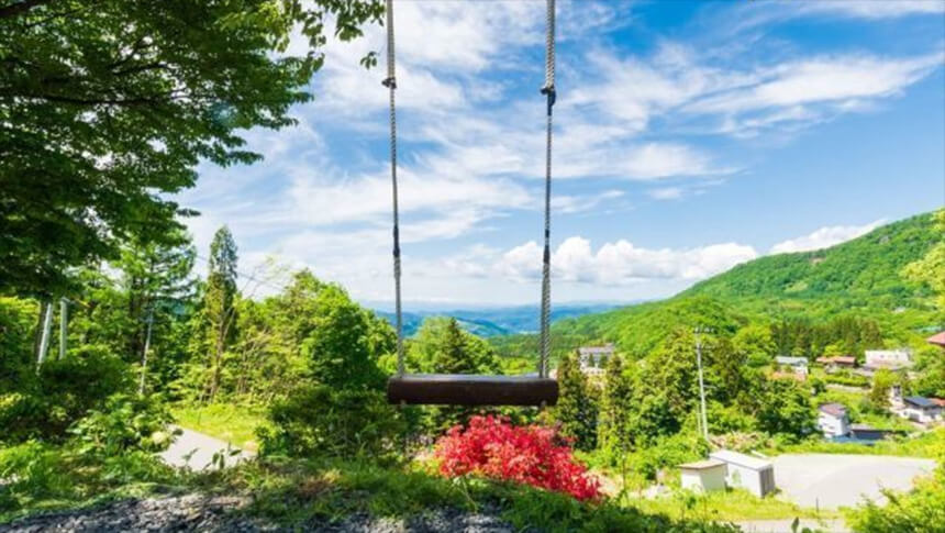 山形県・蔵王温泉にある犬と泊まれる宿「善七の湯」｜高台から蔵王の山並みや街並みを一望できる場所に設置された木製ブランコ。青い空と白い雲、鮮やかな新緑が広がる爽快な風景