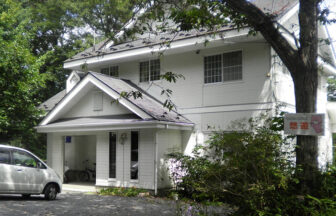 宮城県・蔵王の犬と泊まれる宿「ペンション＆コテージ悠遊」｜緑豊かな木々に囲まれた、白い外壁の2階建てペンション「悠遊」の外観。建物の前には白い車が停まっており、入り口横の木にはピンク色のキャラクターが描かれた看板が設置されている。