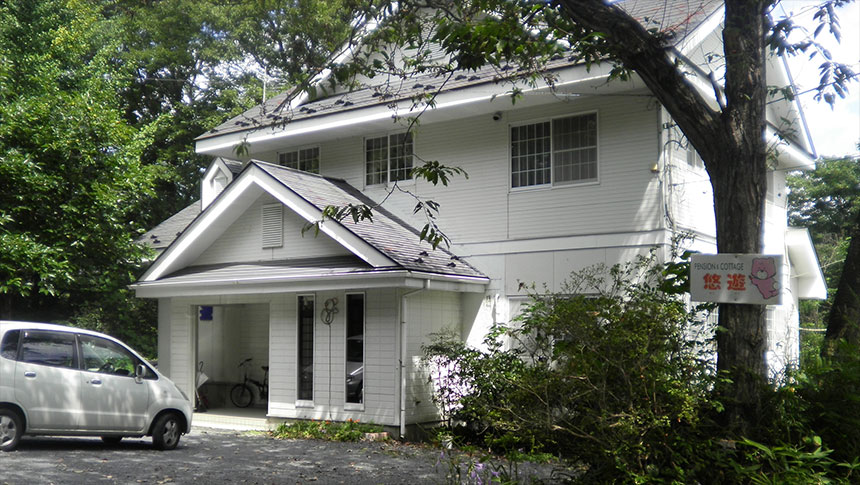 宮城県・蔵王の犬と泊まれる宿「ペンション＆コテージ悠遊」｜緑豊かな木々に囲まれた、白い外壁の2階建てペンション「悠遊」の外観。建物の前には白い車が停まっており、入り口横の木にはピンク色のキャラクターが描かれた看板が設置されている。