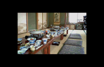 民宿「秋川屋」の食事会場。純和風の広間に、たくさんの料理が載った長机が並んでいる。手前にはカセットコンロに載った大きな鍋、青と白の和食器に盛り付けられた小鉢などがずらりと並び、落ち着いた雰囲気の座敷で楽しむ食事の様子を捉えている