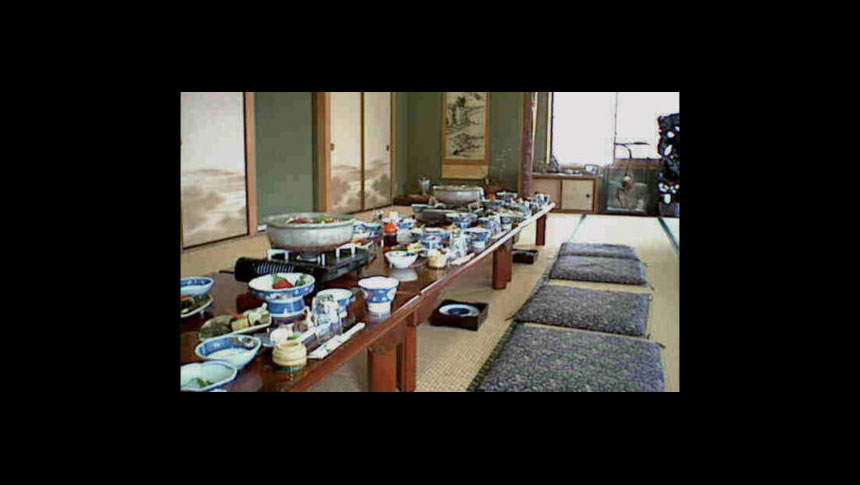 民宿「秋川屋」の食事会場。純和風の広間に、たくさんの料理が載った長机が並んでいる。手前にはカセットコンロに載った大きな鍋、青と白の和食器に盛り付けられた小鉢などがずらりと並び、落ち着いた雰囲気の座敷で楽しむ食事の様子を捉えている