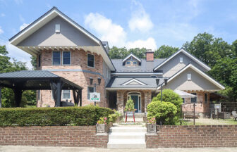 栃木県の那須高原にある犬と泊まれる宿「ザ・ハートフィールドイン」｜洋風の建物の外観写真。煉瓦造りで三角屋根が特徴的。1階にはガレージとテラス、手前には石段と植栽が整えられている。緑豊かな木々に囲まれ、青空が広がっている。