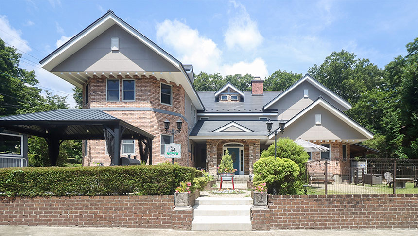 栃木県の那須高原にある犬と泊まれる宿「ザ・ハートフィールドイン」｜洋風の建物の外観写真。煉瓦造りで三角屋根が特徴的。1階にはガレージとテラス、手前には石段と植栽が整えられている。緑豊かな木々に囲まれ、青空が広がっている。