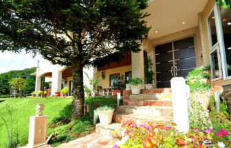 栃木県・那須の犬と泊まれる宿「ザ・ヴィンテージビュー」｜大きな針葉樹の木陰にある建物の入り口。手前にはピンクやオレンジの花が咲き、石積みの階段の脇には白い鉢植えが並んでいます。奥にはモダンな茶色の扉があり、全体的に明るく開放的な雰囲気の外観です