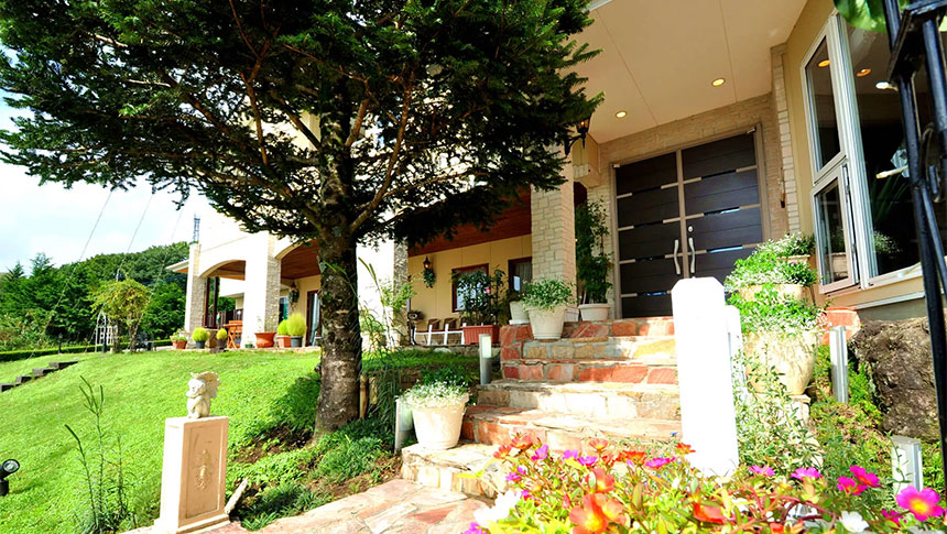 栃木県・那須の犬と泊まれる宿「ザ・ヴィンテージビュー」｜大きな針葉樹の木陰にある建物の入り口。手前にはピンクやオレンジの花が咲き、石積みの階段の脇には白い鉢植えが並んでいます。奥にはモダンな茶色の扉があり、全体的に明るく開放的な雰囲気の外観です