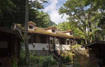 山梨県にある犬と泊まれる宿『クースクース』の外観写真。森の中に佇む、白い壁と3つの屋根窓が特徴的な欧風コテージ。階段付きのテラスがあり、周囲は深い緑の木々に囲まれている。