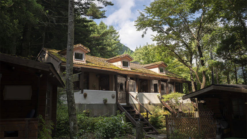 山梨県にある犬と泊まれる宿『クースクース』の外観写真。森の中に佇む、白い壁と3つの屋根窓が特徴的な欧風コテージ。階段付きのテラスがあり、周囲は深い緑の木々に囲まれている。