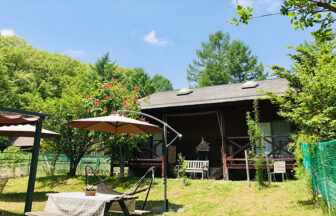 山梨県・清里の犬と泊まれる宿「清里レストラン&コテージ睦(BOKU)」｜緑豊かな森を背景にした、木造のログハウス風コテージの庭。青空の下、テーブル、ベンチ、日よけパラソル、ハンモックが設置され、リラックスできるプライベートな空間が広がっている。