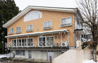 冬の新潟・石打にある犬と泊まれる宿泊施設「石打M・S館」の外観。1階はコンクリート打ちっぱなし、2階以上は温かみのあるオレンジ色の外壁で、大きな三角形の屋根と広々としたバルコニーが特徴。周囲には雪が積もり、隣には高い木々が並んでいる。