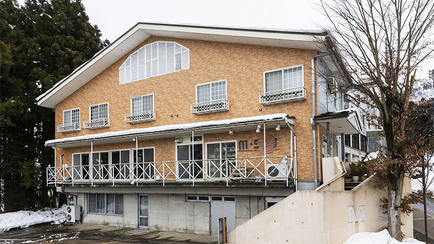 冬の新潟・石打にある犬と泊まれる宿泊施設「石打M・S館」の外観。1階はコンクリート打ちっぱなし、2階以上は温かみのあるオレンジ色の外壁で、大きな三角形の屋根と広々としたバルコニーが特徴。周囲には雪が積もり、隣には高い木々が並んでいる。