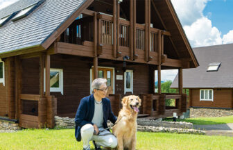 新潟県・当間高原の愛犬と泊まれる宿「ベルナティオ」｜青空の下、木の温もりが感じられる大型のログハウス風コテージの外観。手前の芝生の上では、紺色のジャケットを着た男性が、リードを持った大きなゴールデンレトリバーの横で優しく微笑みながらしゃがんでいます。自然豊かなリゾートでの穏やかなひとときを感じさせる風景です