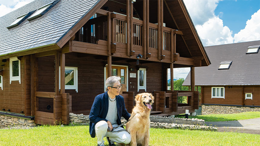 新潟県・当間高原の愛犬と泊まれる宿「ベルナティオ」｜青空の下、木の温もりが感じられる大型のログハウス風コテージの外観。手前の芝生の上では、紺色のジャケットを着た男性が、リードを持った大きなゴールデンレトリバーの横で優しく微笑みながらしゃがんでいます。自然豊かなリゾートでの穏やかなひとときを感じさせる風景です