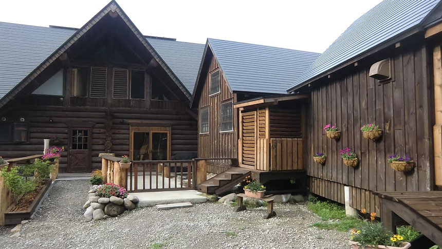 長野県にある犬と泊まれる宿『コテージ・ログハウスGOO』の外観写真。重厚な丸太造りのログハウスと木造コテージが並び、建物の壁や入り口付近には色鮮やかな花が飾られたプランターやハンギングバスケットが並んでいる。