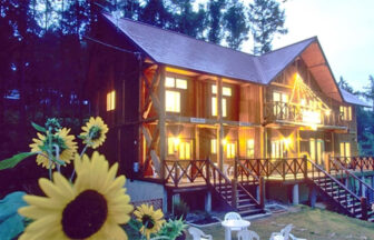 長野県・タングラム斑尾の犬と泊まれる宿「高原のログP.カンタベリー」｜森を背景にした木造2階建てペンションの夕景。窓やベランダから漏れる暖色の光が建物を温かく照らしています。手前左側にはひまわりの花が大きく写り、庭には白いテーブルと椅子のセット。ペンションへの階段とベランダは木製で、落ち着いた雰囲気が漂っています