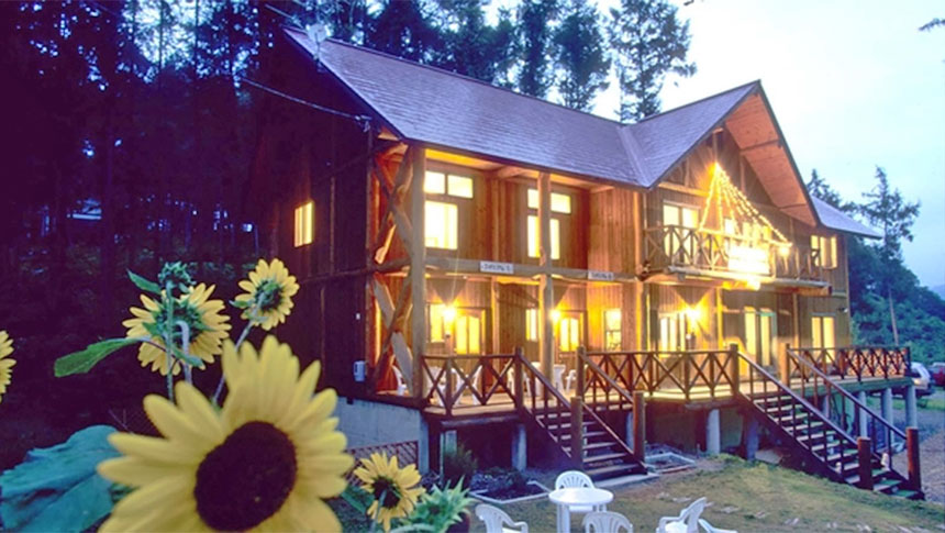 長野県・タングラム斑尾の犬と泊まれる宿「高原のログP.カンタベリー」｜森を背景にした木造2階建てペンションの夕景。窓やベランダから漏れる暖色の光が建物を温かく照らしています。手前左側にはひまわりの花が大きく写り、庭には白いテーブルと椅子のセット。ペンションへの階段とベランダは木製で、落ち着いた雰囲気が漂っています