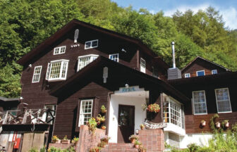 深い緑の森を背に建つ、ダークブラウンの外壁が特徴的な3階建ての山小屋|長野県・乗鞍高原の犬と泊まれる宿「ボエティカル」の外観。白い窓枠やポーチのレンガ造りがアクセントとなり、エントランスには鉢植えが並んでいる