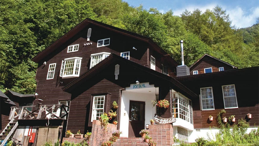 深い緑の森を背に建つ、ダークブラウンの外壁が特徴的な3階建ての山小屋|長野県・乗鞍高原の犬と泊まれる宿「ボエティカル」の外観。白い窓枠やポーチのレンガ造りがアクセントとなり、エントランスには鉢植えが並んでいる