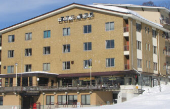 長野・栂池高原に佇む犬と泊まれる宿「びわ池ホテル」の外観。ベージュの建物は雪景色に映え、屋根近くの看板が目印。目の前に広がる真っ白な雪原は、スキーシーズンにはゲレンデ直結の利便性を感じさせる。1階の大きな窓からは、雪景色を眺めながらくつろげる様子が窺える。