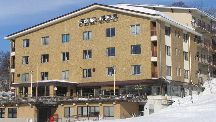 長野・栂池高原に佇む犬と泊まれる宿「びわ池ホテル」の外観。ベージュの建物は雪景色に映え、屋根近くの看板が目印。目の前に広がる真っ白な雪原は、スキーシーズンにはゲレンデ直結の利便性を感じさせる。1階の大きな窓からは、雪景色を眺めながらくつろげる様子が窺える。