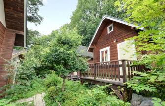 長野県・信州の犬と泊まれる宿「信州愛和の森ホテル」｜森の中に佇む、木製の壁が特徴的なログハウス風のコテージ。手前には白い手すりのあるテラス席があり、緑豊かな木々や植物に囲まれている。左側にも木製の壁の建物が一部見え、石畳のような小道が続いている。