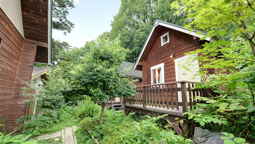 長野県・信州の犬と泊まれる宿「信州愛和の森ホテル」｜森の中に佇む、木製の壁が特徴的なログハウス風のコテージ。手前には白い手すりのあるテラス席があり、緑豊かな木々や植物に囲まれている。左側にも木製の壁の建物が一部見え、石畳のような小道が続いている。