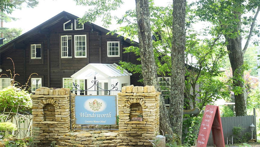 長野県・八ヶ岳中央高原　原村の犬と泊まれる宿「Wandsworth」｜石造りの門柱と『Wandsworth』の看板が印象的な、木々に囲まれた英国風コテージの外観。黒い木壁に白い窓枠が映えるシックな建物