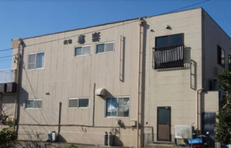 愛知県南知多の民宿 喜楽（きらく）の外観写真。ベージュのサイディング外壁に「民宿 喜楽」と黒い文字で書かれている。2階建ての建物で、左側には小さなバルコニーがあり、1階部分にはプロパンガスのボンベや入り口のドアが見える。快晴の空を背景にした、静かで落ち着いた佇まい